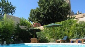 A clear blue swimming pool with lounge chairs and umbrellas, surrounded by lush greenery and a castle-like building in the background. - Grand old Villa with pool in the heart  of  Capestang town (on the Canal du Midi) (Capestang)