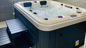 Ein Badezimmer mit einer großen, dunkel gefärbten Whirlwanne, einem kleinen Holzschemel und einem Fenster mit Tageslicht.