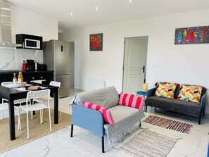 Ein modernes Wohnzimmer mit Sofa, Esstisch und Stühlen, Küchenzeile mit Mikrowelle und Kaffeemaschine sowie einer Tür zu einem weiteren Raum.