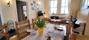 A well-lit living room with a dining area, a sofa, a TV, and a table set for two with a vase of flowers.