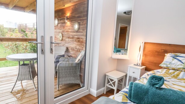 A bedroom with a sliding glass door leading to a deck, a bed with a patterned duvet, a bedside table with a lamp, and a small white nightstand.