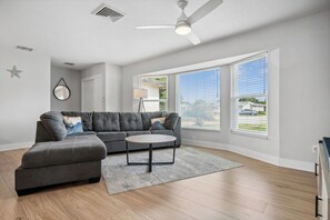 Ein modernes Wohnzimmer mit einem Ecksofa, einem runden Couchtisch und großen Fenstern mit Jalousien.