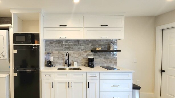 A modern kitchen with white cabinets, a black refrigerator, a microwave, a coffee maker, and a tiled backsplash. - #79 The Summer Loft by Joyful Moments Vacations (Clearwater)