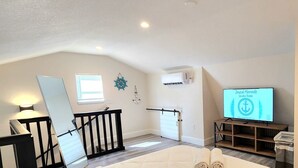 A living room with a TV displaying 'Joyful Moments Vacation Planner', a wooden TV stand, a white door, and a staircase with a black railing. - #79 The Summer Loft by Joyful Moments Vacations (Clearwater)