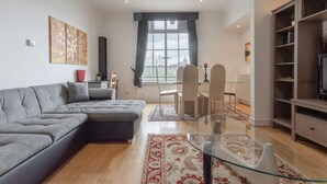 A modern living room with a grey sectional sofa, a glass coffee table, and a dining area with a table and chairs. - Large & Central 2BD Flat - Tower Hill (London)