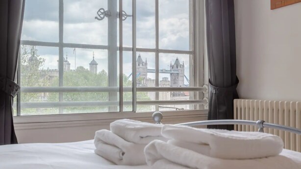A hotel room with a bed, a window with a view of a bridge and buildings, and a radiator. - Large & Central 2BD Flat - Tower Hill (London)