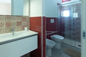 Een badkamer met een wit wastafelmeubel, twee toiletten en een douchegedeelte met betegelde muren.