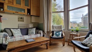 Una sala de estar acogedora con un sofá, una mesa de centro y una silla. Se ve una ventana con vistas a edificios y árboles.