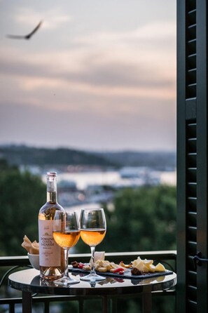 A table with a bottle of wine and two glasses, set against a scenic view of a body of water and hills. - Penthouse Suite Sea View (Corfu)