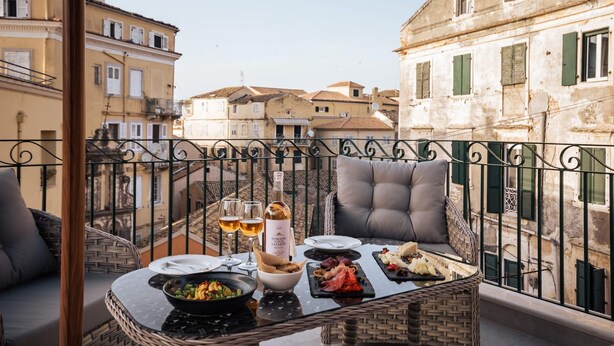 A balcony with a dining table set for two, featuring a bottle of wine, glasses, and a variety of food. - Penthouse Suite Sea View (Corfu)