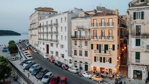 A multi-story building with balconies, a street with parked cars, and a body of water in the background. - Penthouse Suite Sea View (Corfu)