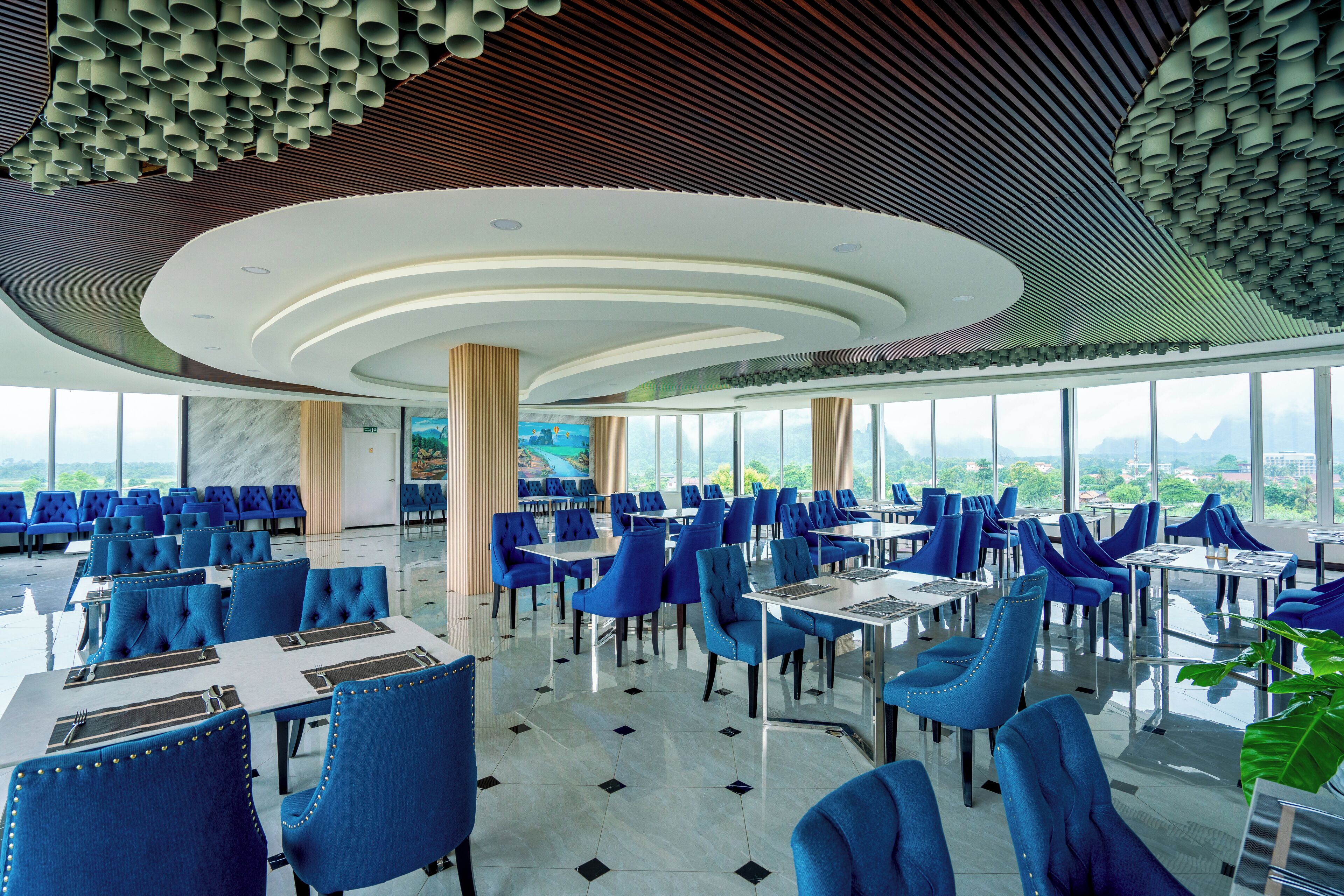 A modern restaurant with blue upholstered chairs and tables, large windows offering a city view, and a unique ceiling design with cylindrical elements.