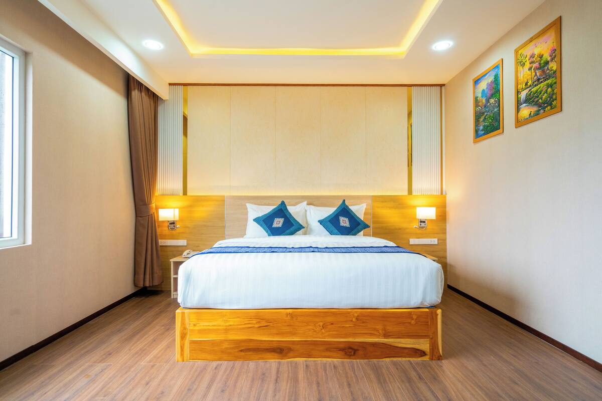 Ein Hotelzimmer mit einem hölzernen Bett, zwei Nachttischlampen, einem fenster mit Vorhängen und einem Gemälde an der Wand.