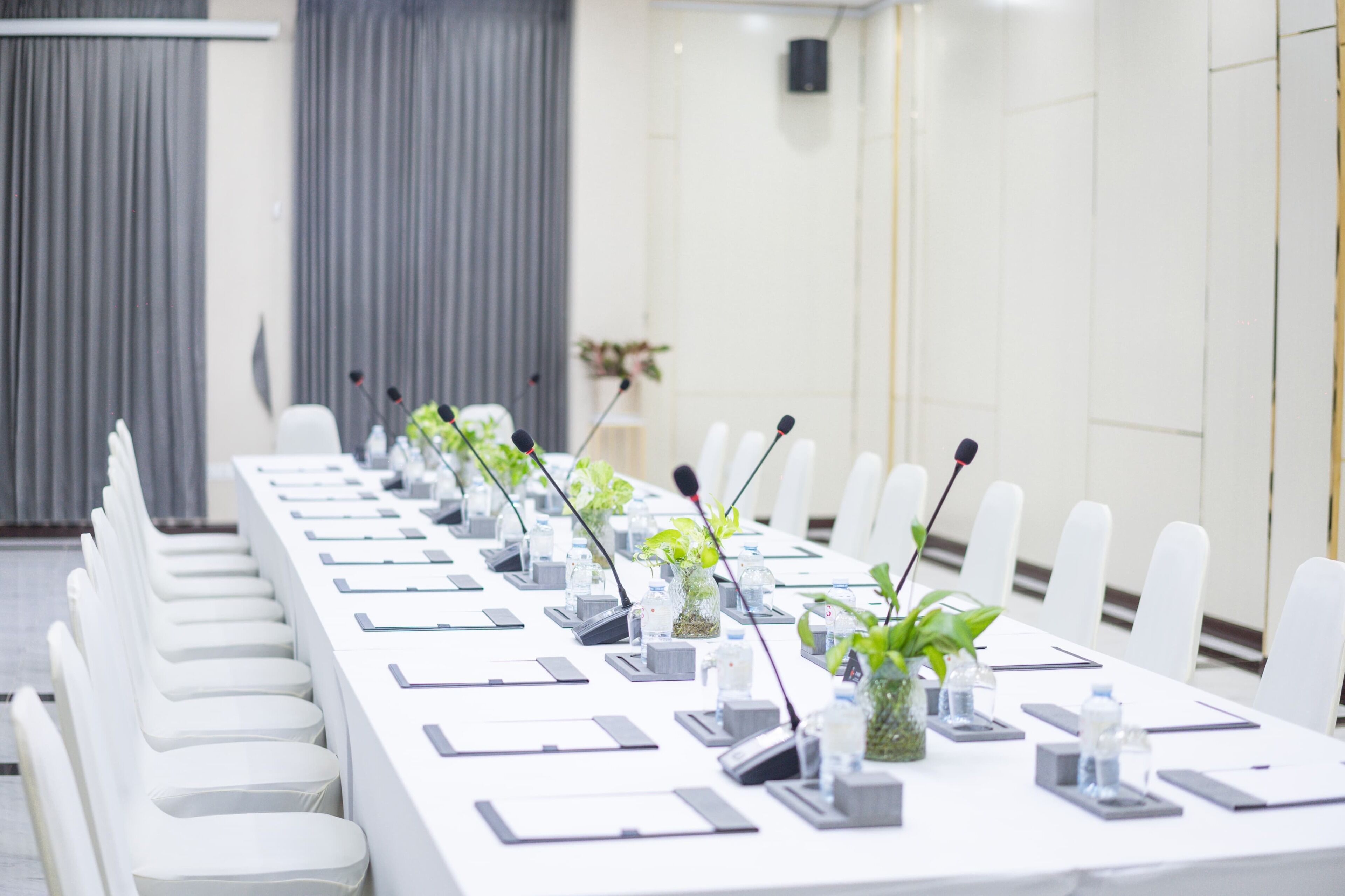 Un salón de conferencias con una larga mesa, sillas, micrófonos y pequeñas macetas.
