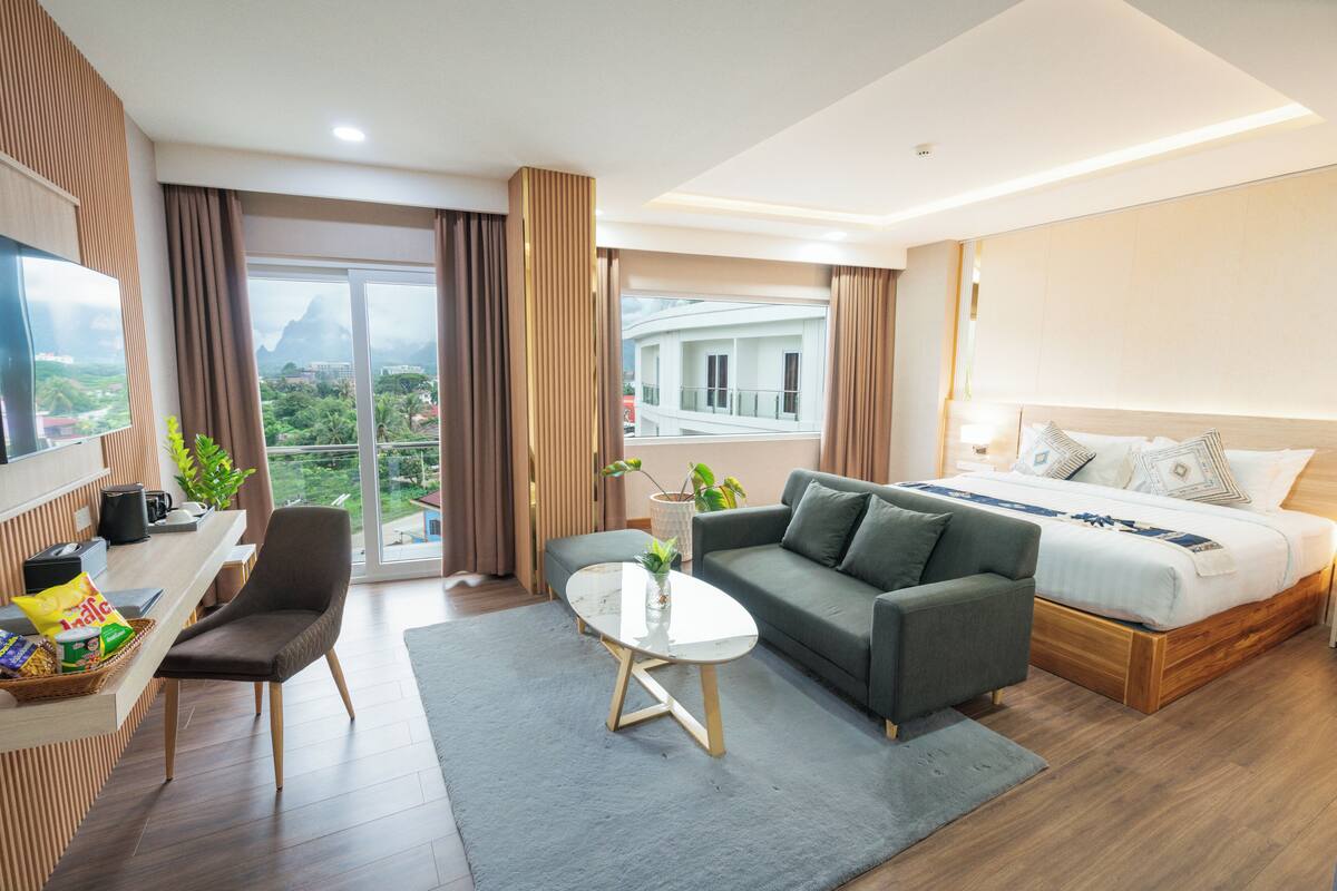 Ein modernes Hotelzimmer mit einer Couch, einem Bett, einer Essecke mit Tisch und Stühlen sowie einem großen Fenster mit Blick auf die Stadt.