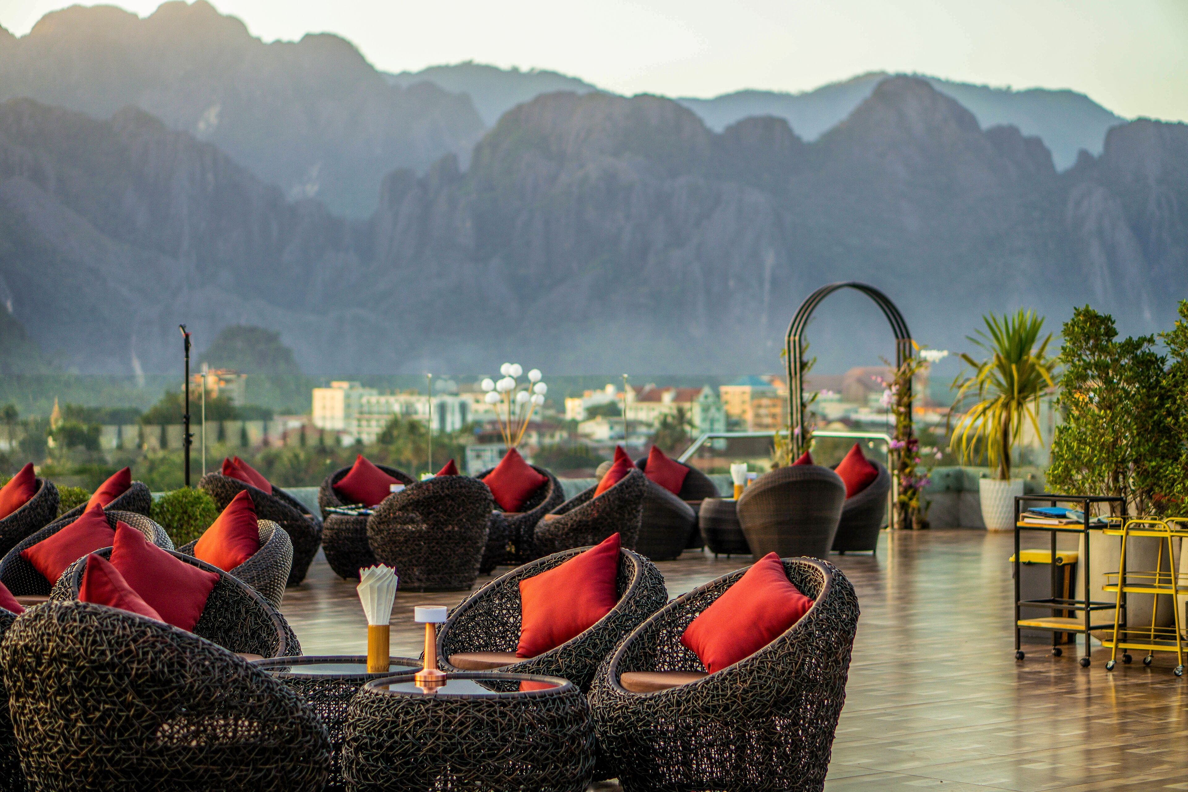 Terraza en la azotea con sillas de mimbre y cojines rojos, con vistas a un paisaje urbano y montañas.