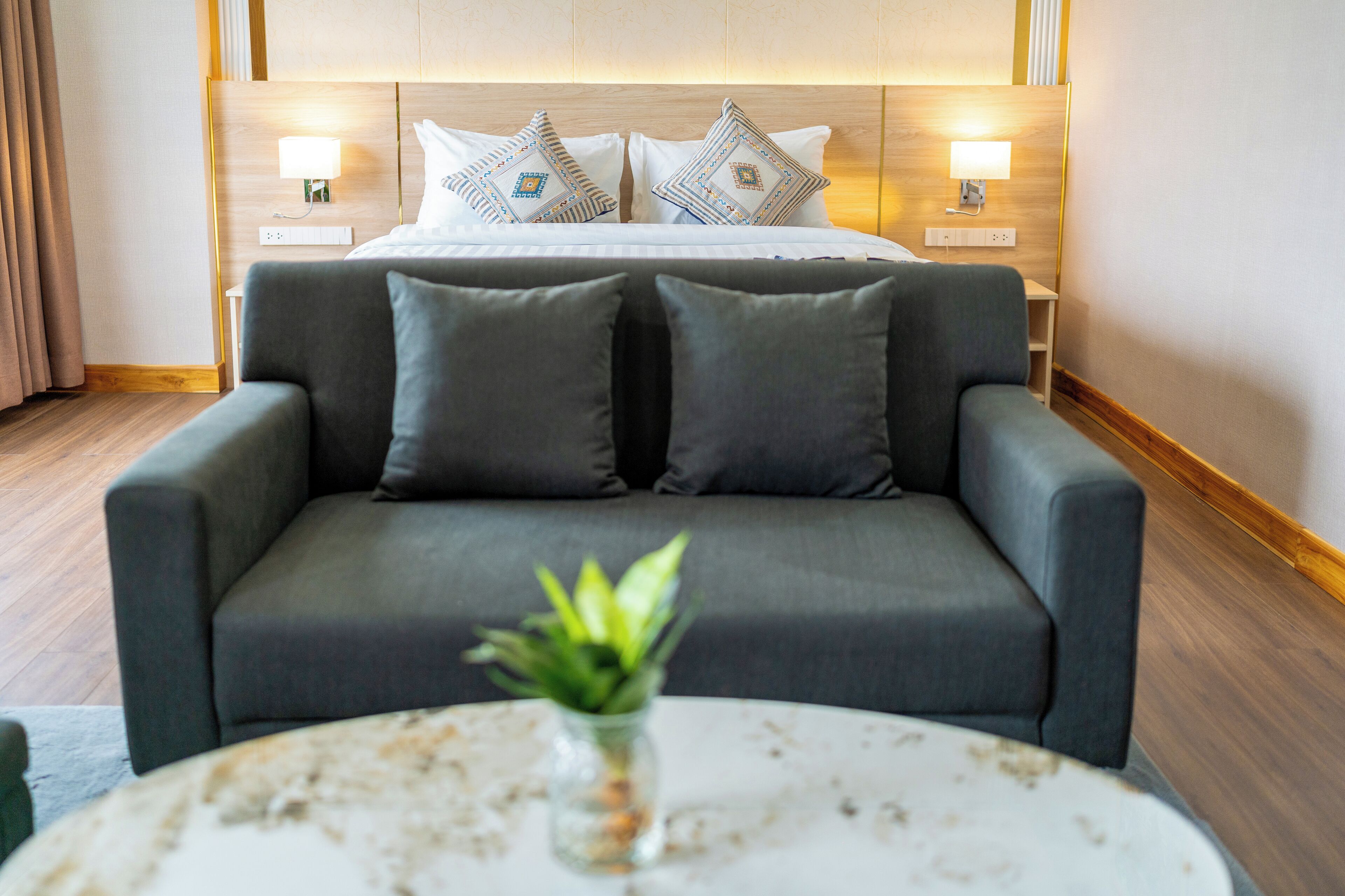Habitación de hotel moderna con una cama grande, dos lámparas de noche, un sofá gris y una mesa redonda con una planta.
