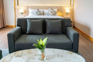 Habitación de hotel moderna con una cama grande, dos lámparas de noche, un sofá gris y una mesa redonda con una planta.