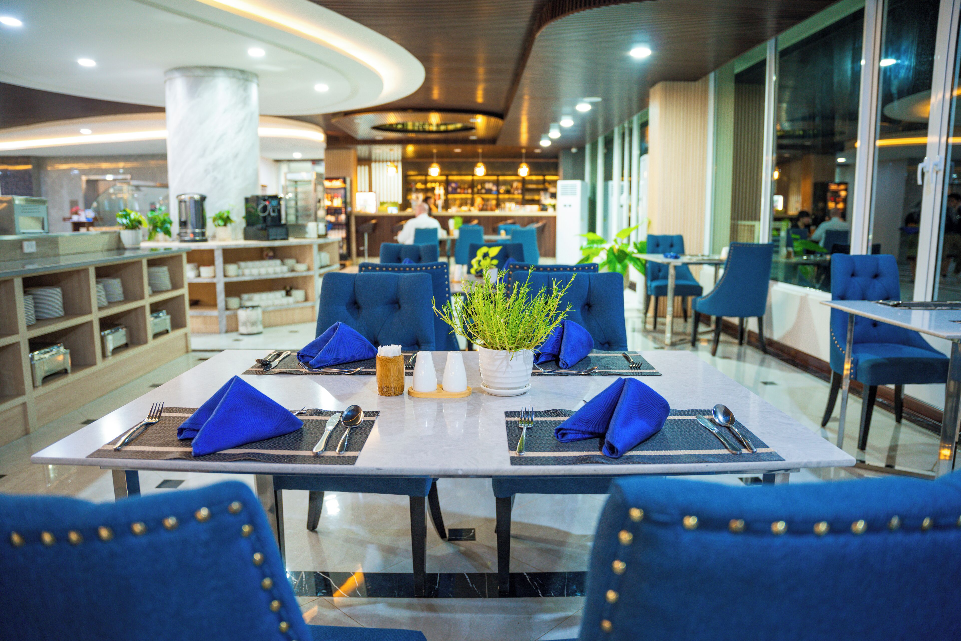 A restaurant with a modern interior, featuring a marble column, blue upholstered chairs, and a central table with a potted plant.