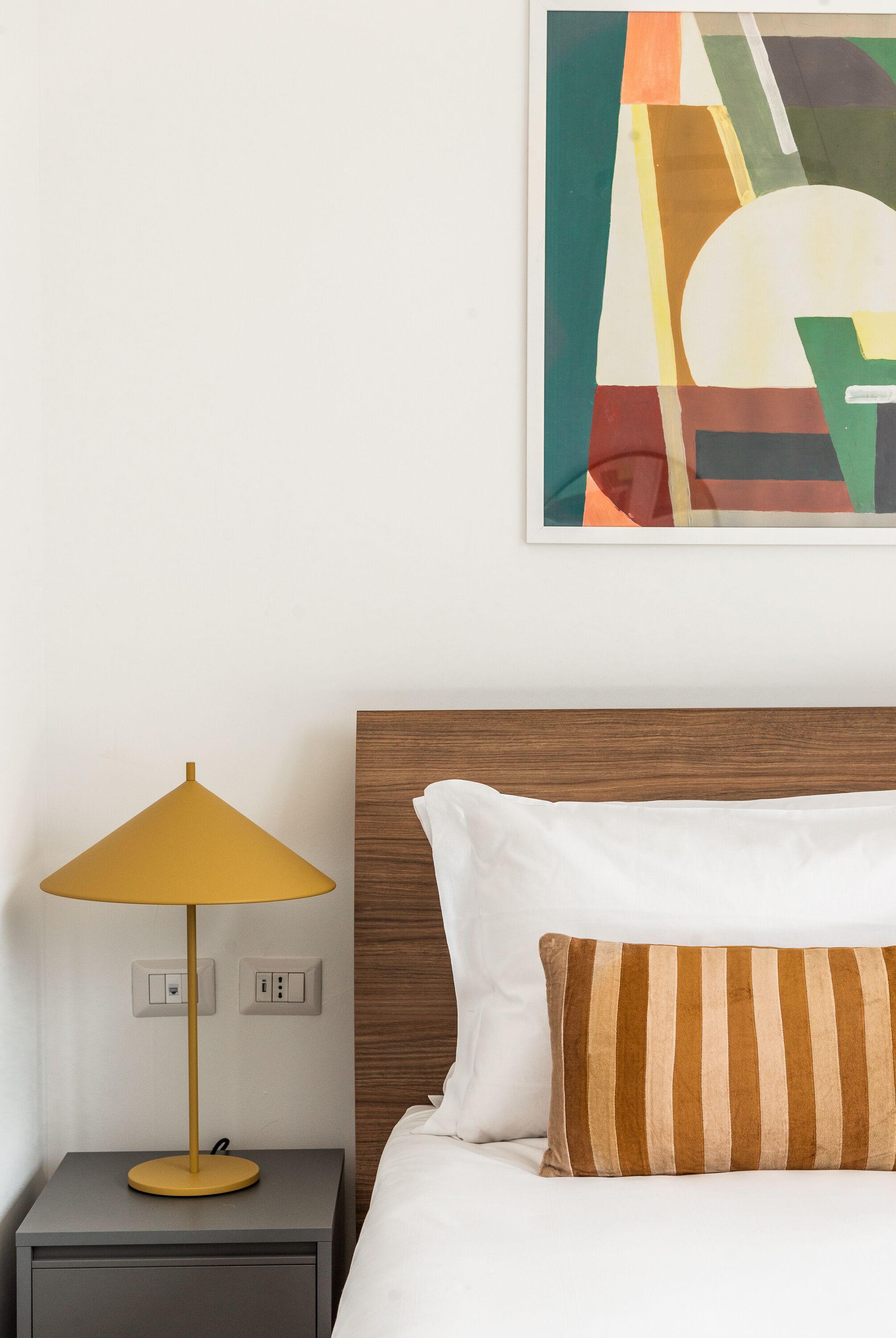 A neatly made bed with a striped pillow, a bedside table with a yellow lamp, and a colorful abstract painting on the wall.