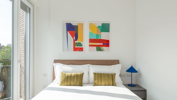 Un dormitorio moderno con una cama grande, dos almohadas, una mesita de noche con una lámpara y arte abstracto en la pared.