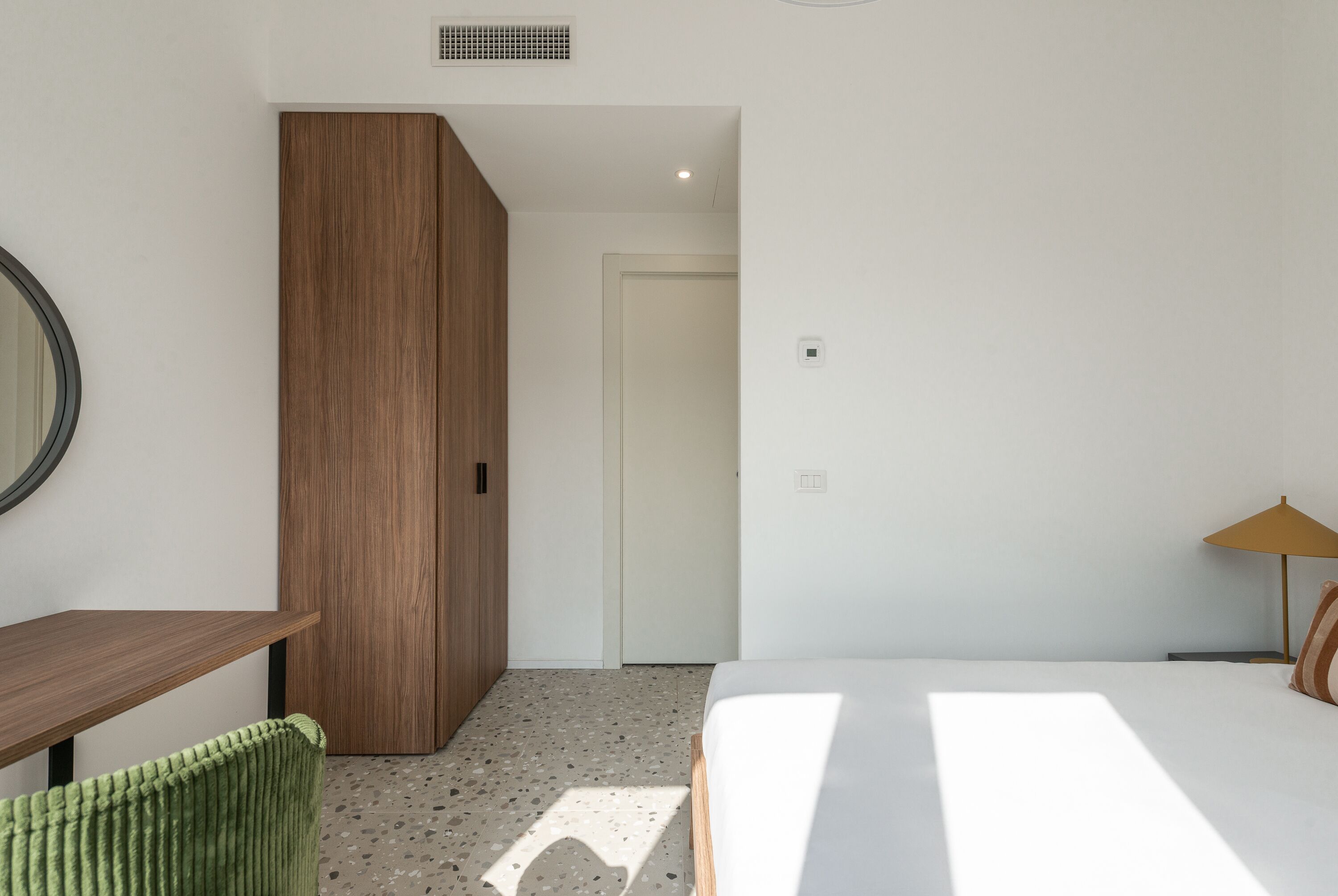 A modern bedroom with a wooden wardrobe, a green chair, a bed with white bedding, and a small table.