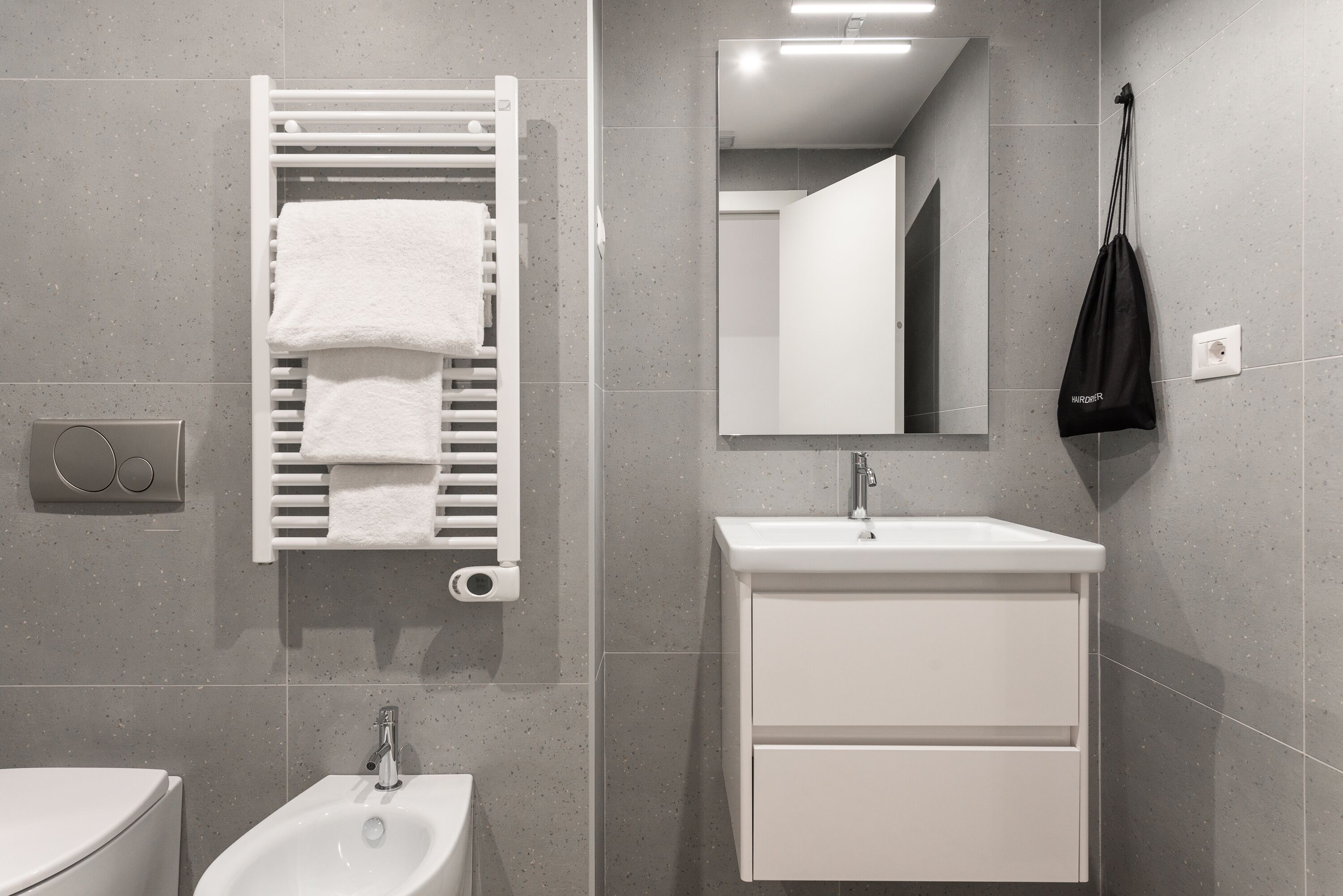 A modern bathroom with a white sink, a bidet, a towel rack with two white towels, a mirror, and a black bag hanging on the wall.