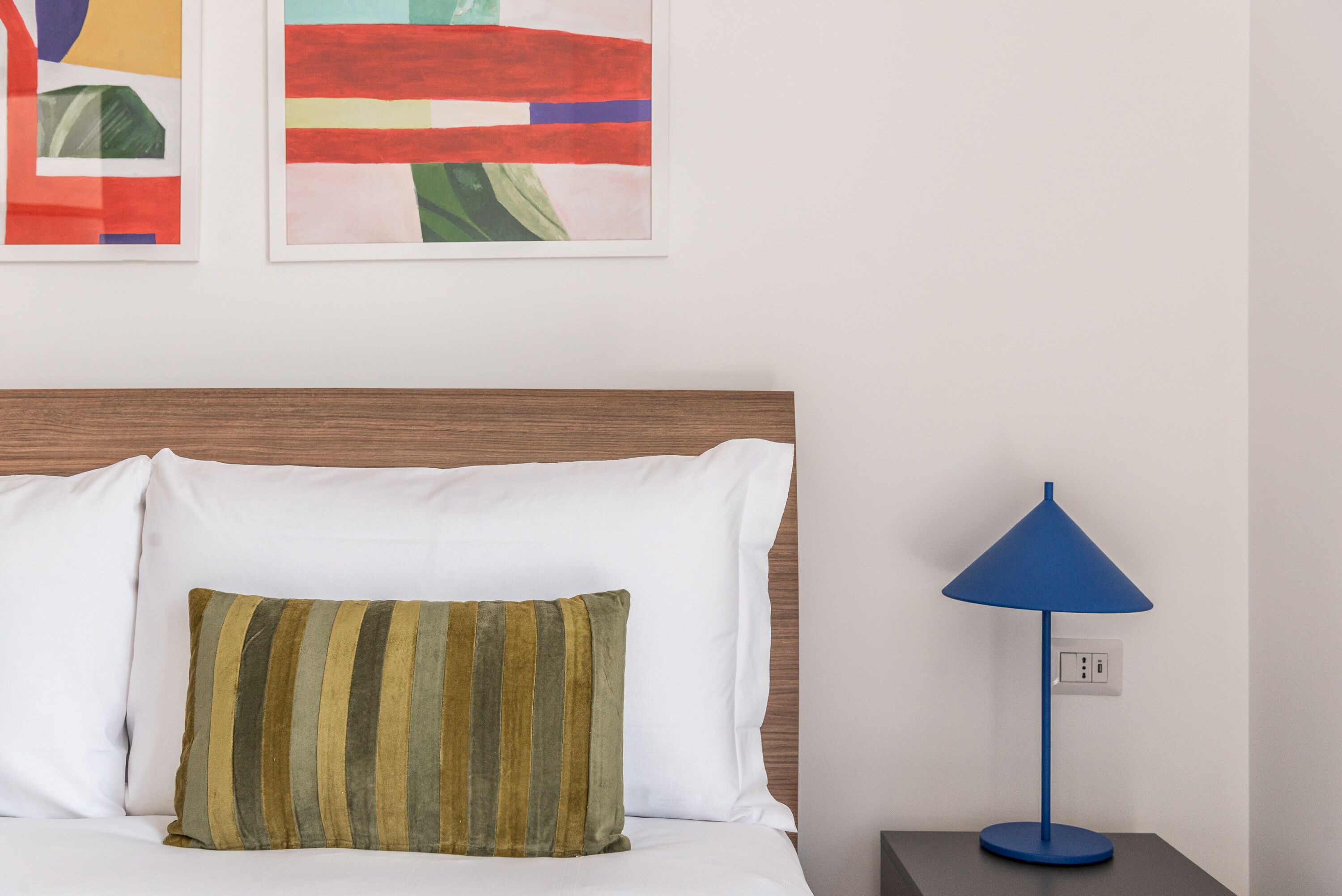 A neatly made bed with white and striped pillows, a blue lamp on a bedside table, and abstract art on the wall.