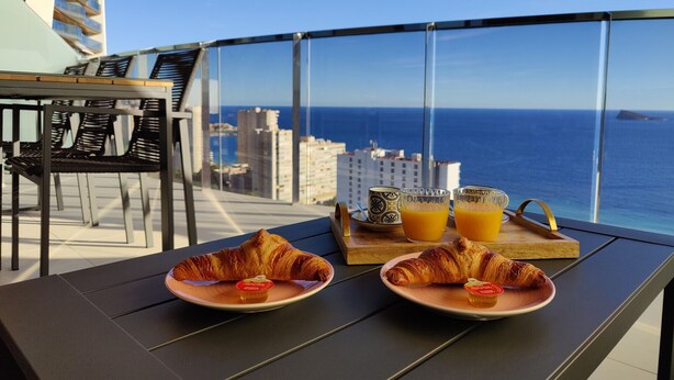 Un balcón con dos platos de cruasanes y jugo de naranja, una bandeja de madera, y vistas al océano y edificios.