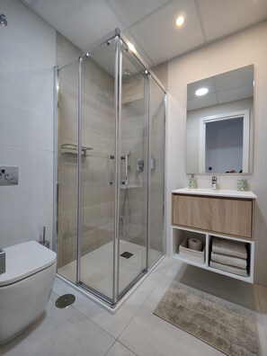 A modern bathroom with a glass-enclosed shower, a white toilet, a wooden vanity with a mirror, and a beige rug. - SUNSET WAVES superb apartments (Benidorm)