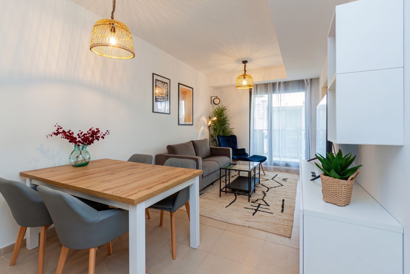 Ein modernes Wohnzimmer mit einem Holzesstisch, grauen Stühlen, einer Couch und einem Couchtisch. Zwei Pendelleuchten, ein Blumentopf und ein Teppich liegen auf dem Boden.