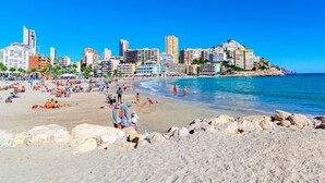 Una platja amb gent que pren el sol i nedant, una riba rocosa i un paisatge urbà amb edificis alts a la part de darrere.