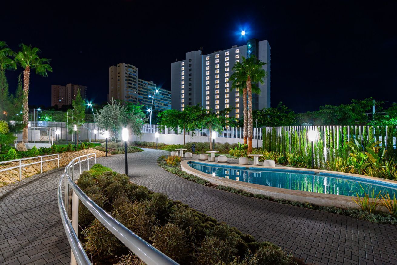 A nighttime view of a landscaped area with a swimming pool, palm trees, and a pathway leading to a building with illuminated windows.