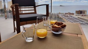 Un balcon avec vue sur la mer, une chaise en bois et une table sur laquelle se trouvent un verre de jus d’orange, un verre d’eau et un bol de croissants.