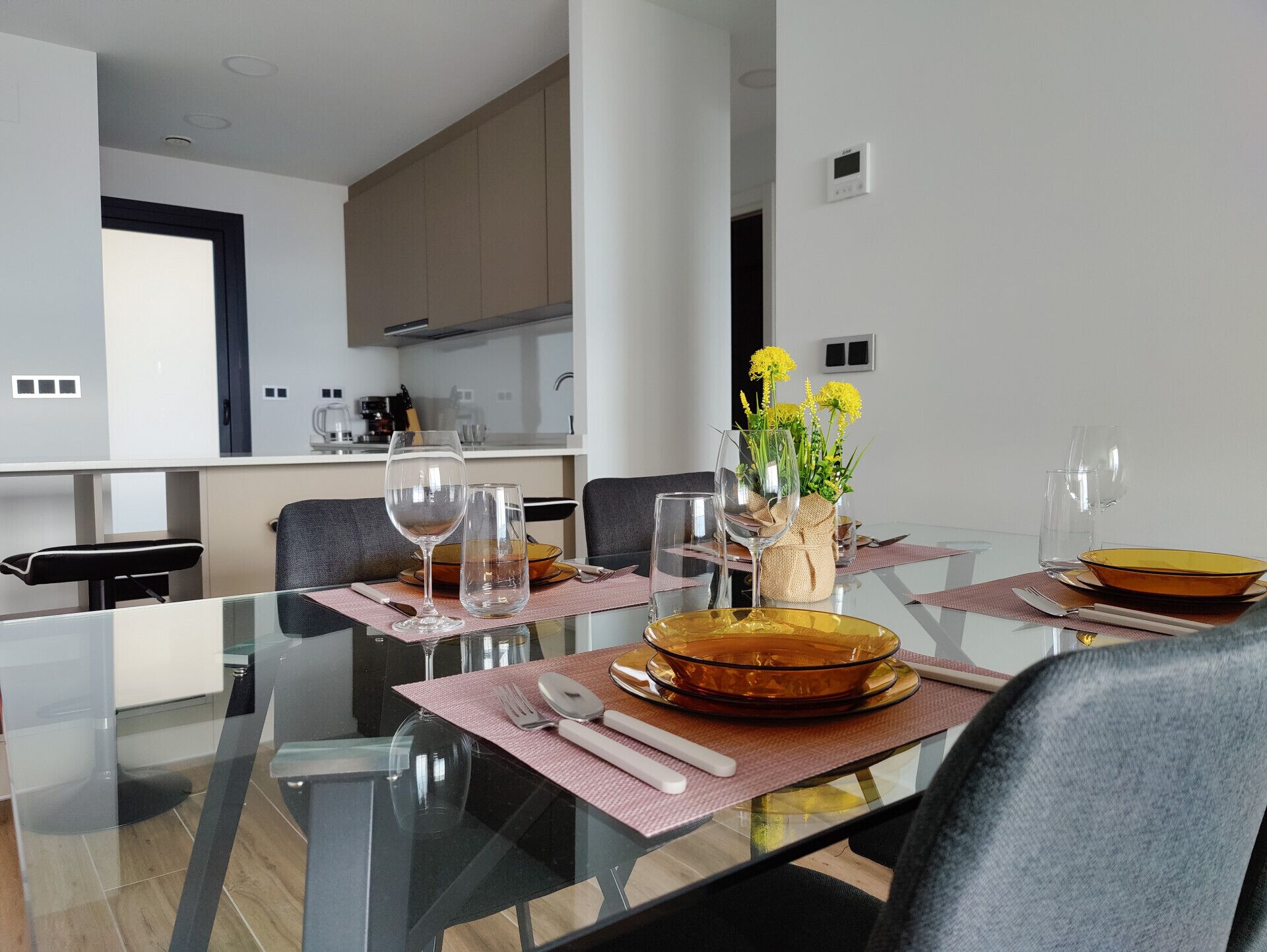 Un espace repas moderne avec une table en verre, de la verrerie transparente et un vase rempli de fleurs jaunes.