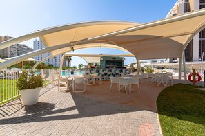 An outdoor patio area with white tables and chairs, a bar, and a pool in the background. - SUNSET WAVES apARTs by RB (Benidorm)