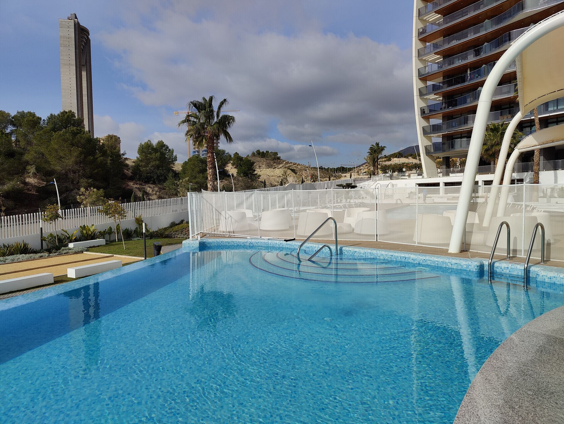 Una piscina con agua azul cristalina, rodeada por una cerca blanca y palmeras.