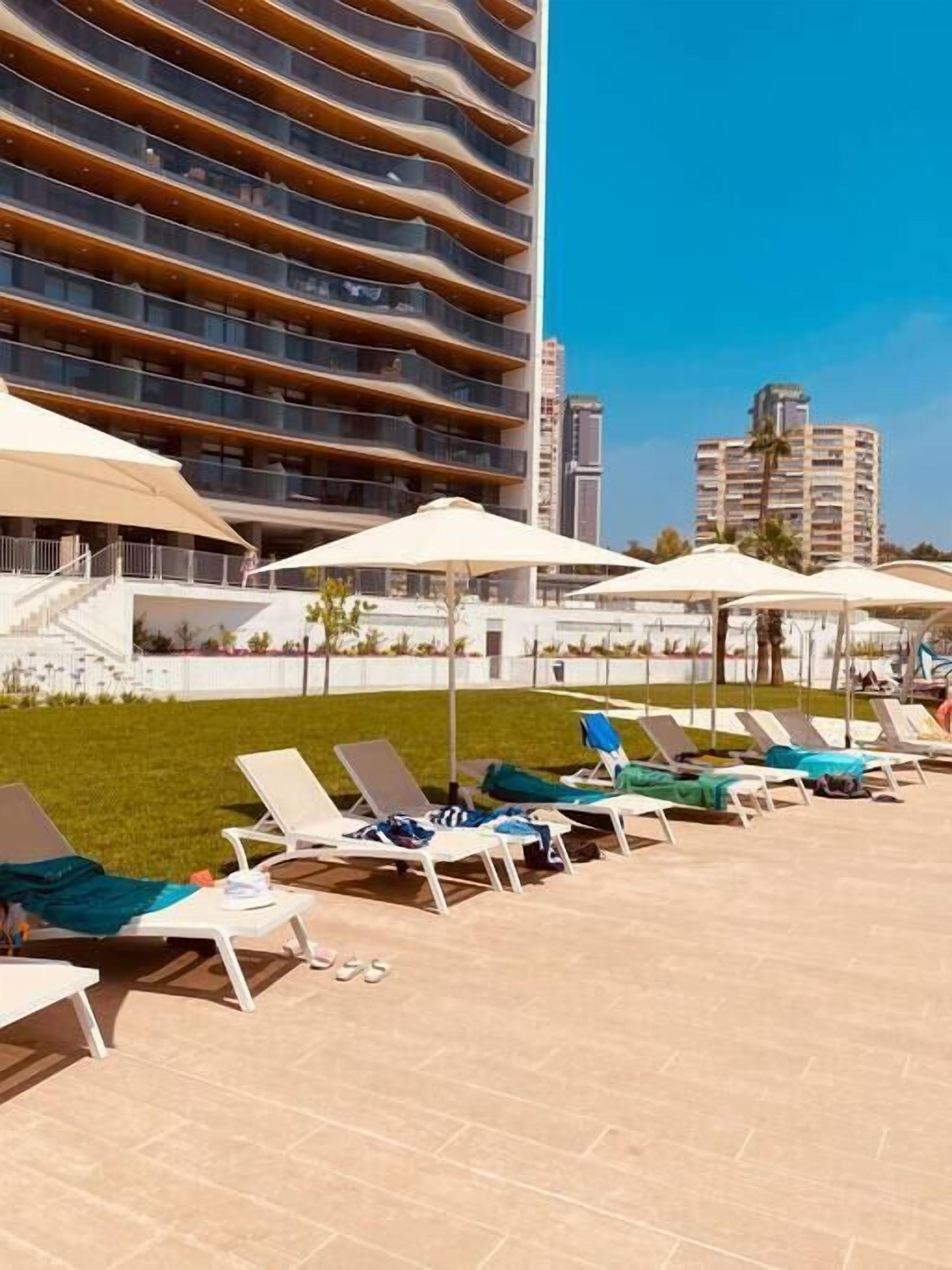 A rooftop pool area with lounge chairs and umbrellas, surrounded by a modern building with balconies.