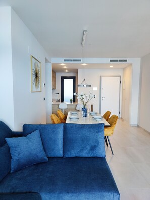 A modern living room with a blue sofa, a dining table set with plates and glasses, and a kitchen in the background.