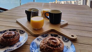 Ett träbord med frukosttillbehör, inklusive bakverk och apelsinjuice, på en balkong med utsikt över havet och byggnaderna.