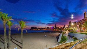 Un paisaje urbano costero al atardecer, con una playa, palmeras y edificios iluminados.