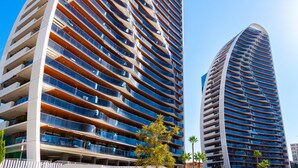 Dos edificios modernos altos con varios balcones, cielo despejado y algo de vegetación.
