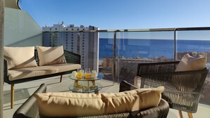 Un balcón con sofá y sillones, una mesa de centro de vidrio con jugo de naranja y vista al mar y edificios.