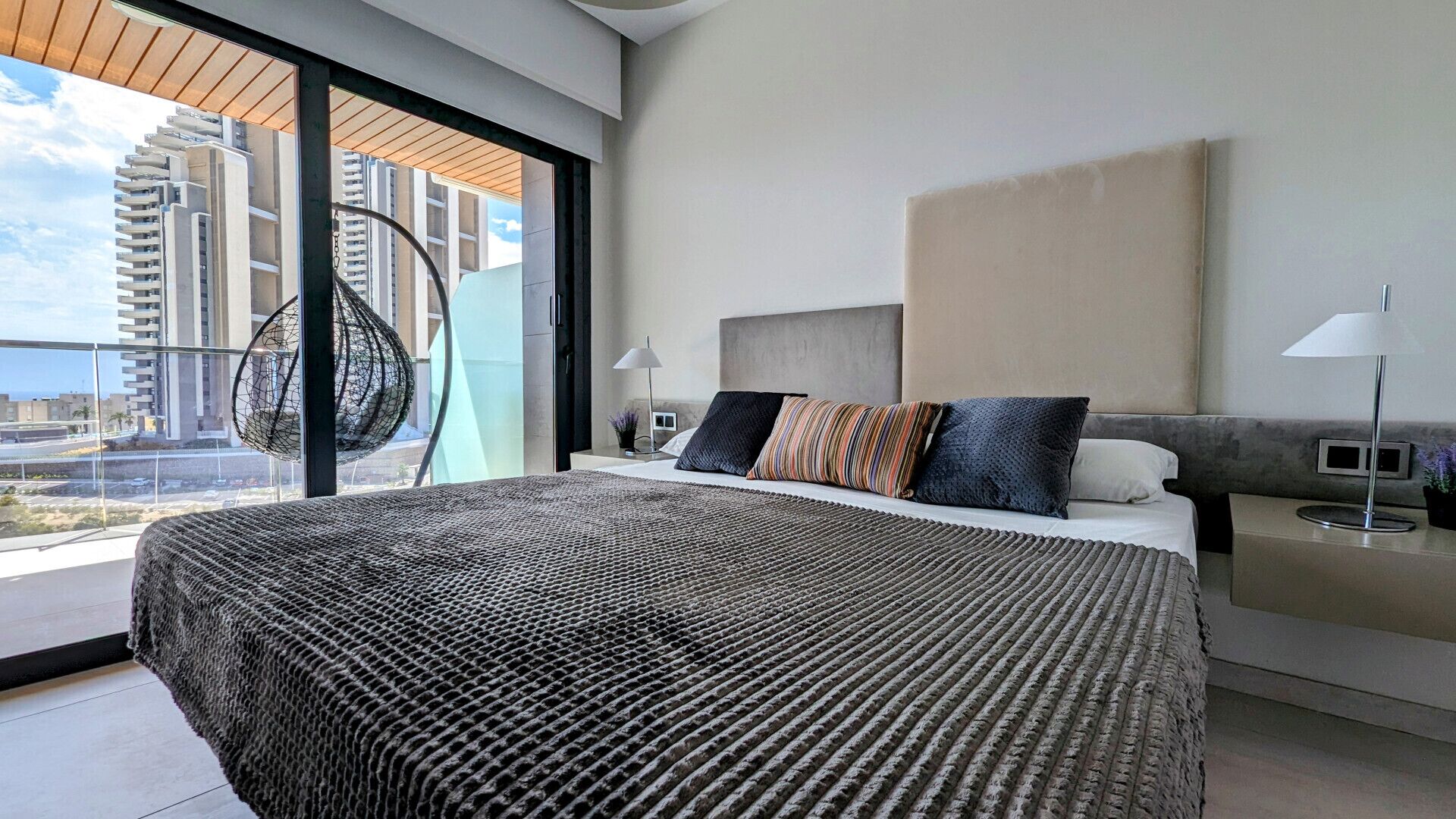 Quarto de hotel moderno com uma cama grande, vista para a cidade e varanda com cadeira suspensa.