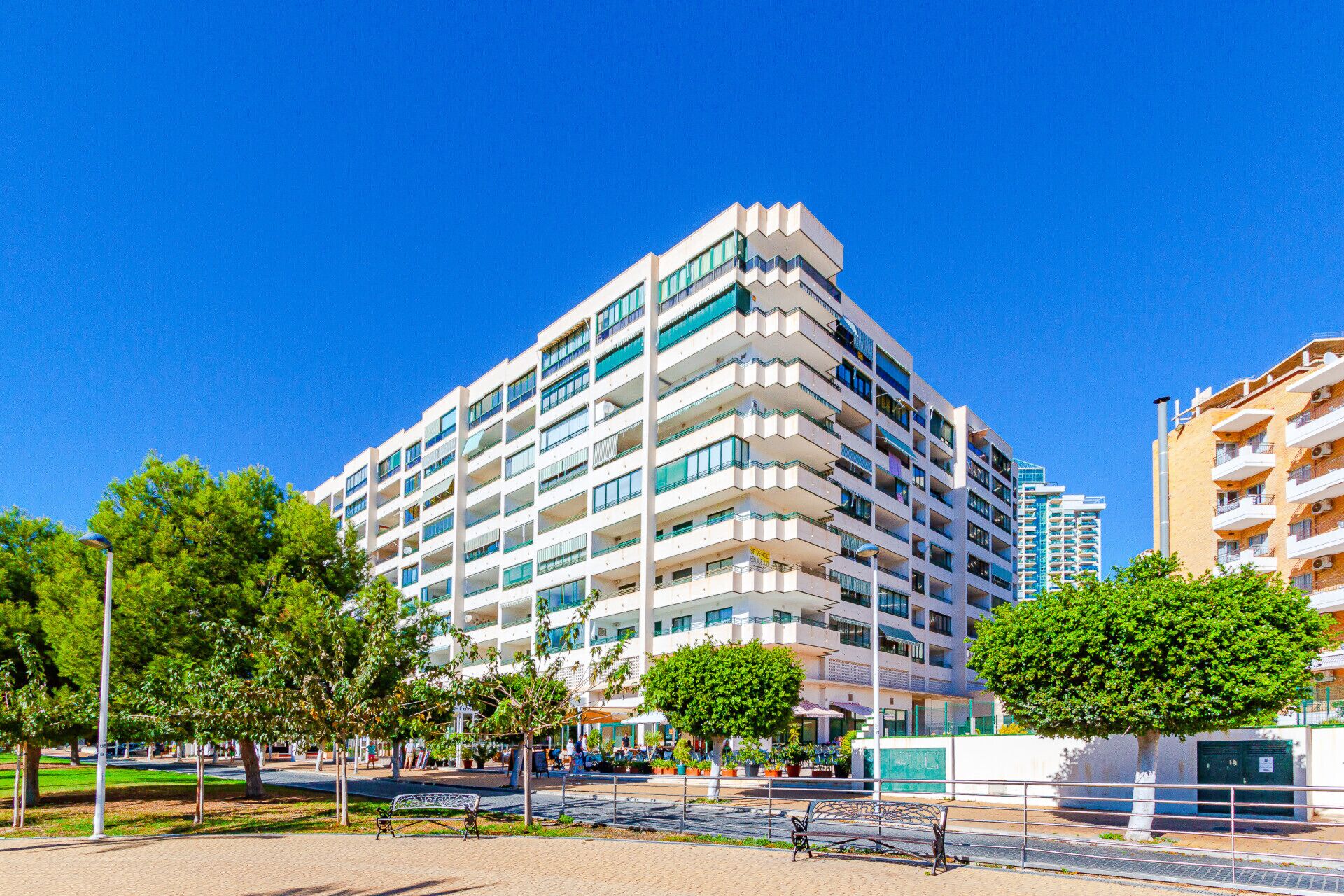 A multi-story hotel with balconies, a paved area with benches, and trees.