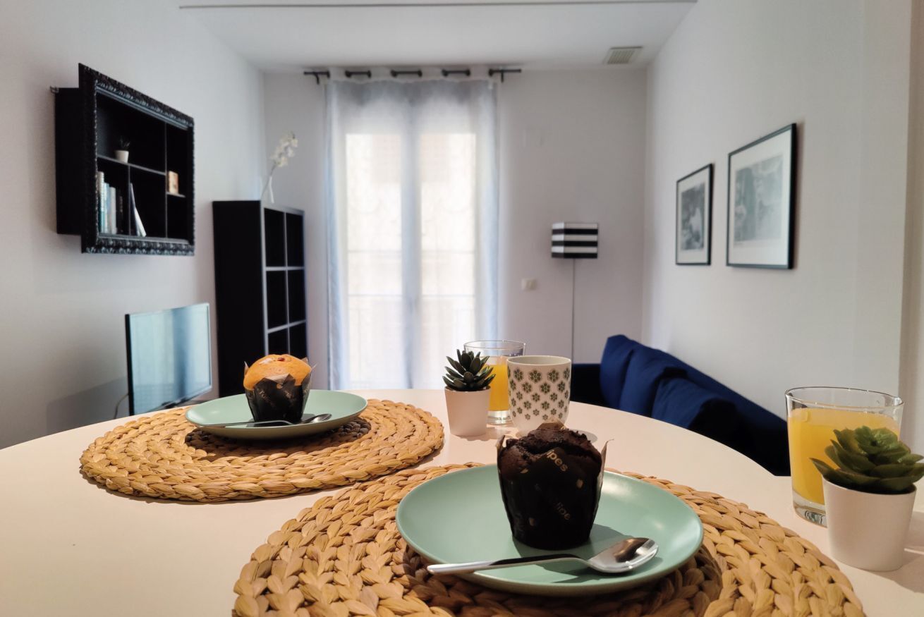 Una sala de estar moderna con una mesa redonda, dos individuales tejidos, un plato verde con un muffin, un vaso de jugo de naranja y una pequeña planta en maceta.