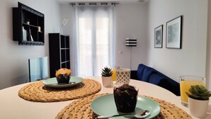 Un salon moderne avec une table ronde, deux sets de table en fibres naturelles, une assiette verte contenant un muffin, un verre de jus d’orange et une petite plante en pot.