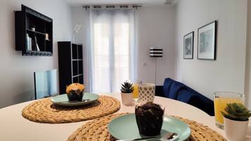 Una sala de estar moderna con una mesa redonda, dos individuales tejidos, un plato verde con un muffin, un vaso de jugo de naranja y una pequeña planta en maceta.