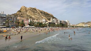 Une plage où l’on voit des gens nager et bronzer, une grande colline avec des bâtiments en arrière-plan et un ciel dégagé.