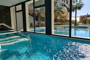 A swimming pool with clear blue water, a metal ladder, and large windows offering views of palm trees and buildings. - ALLONBAY spa & beach house (Villajoyosa)